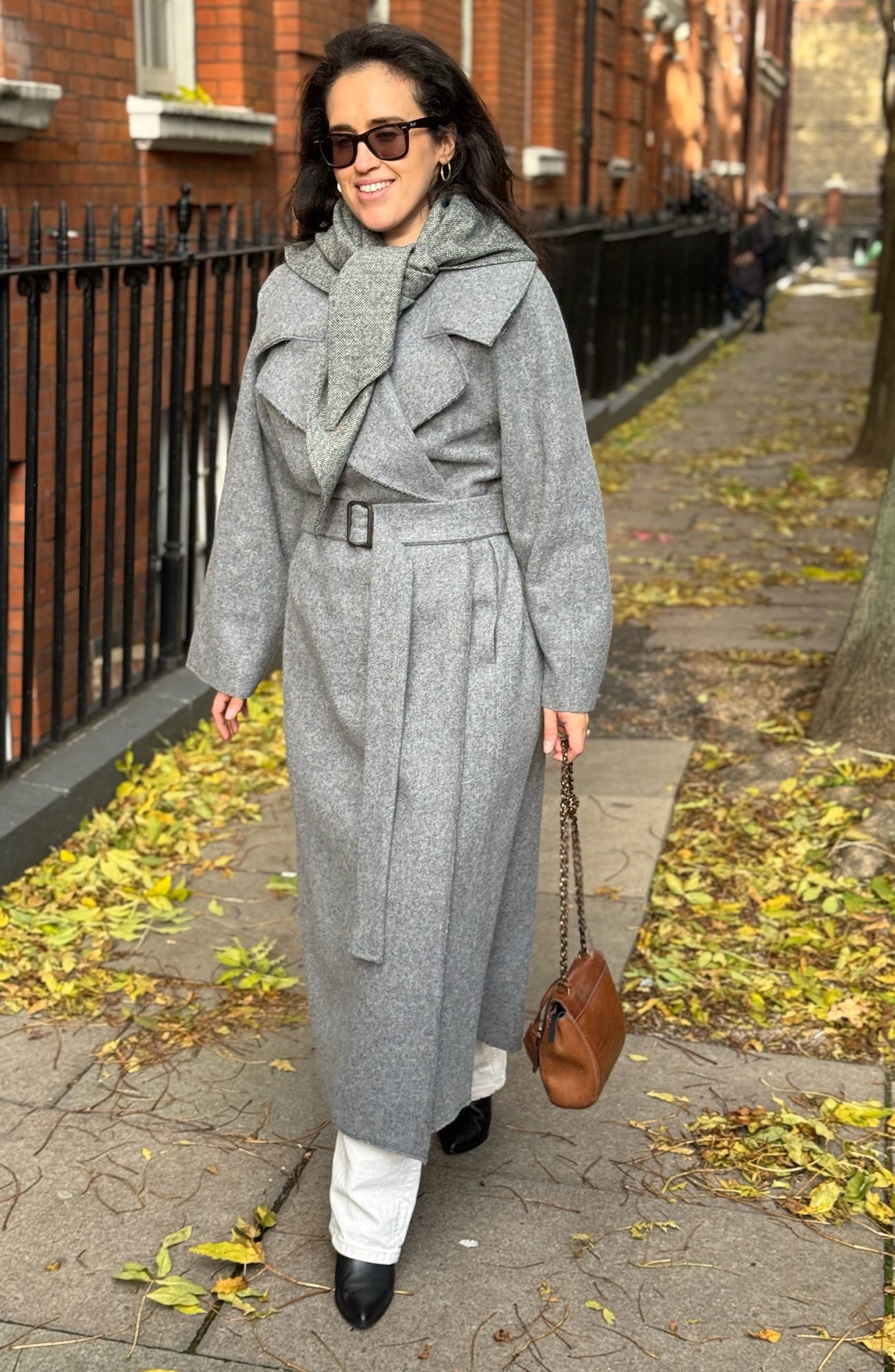 Woman in a gray coat walking on a sidewalk with autumn leaves.
